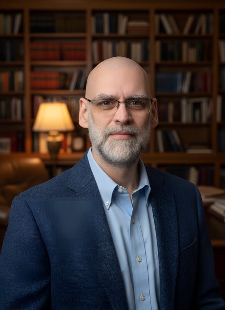 Raymond Brunell, a bearded man in glasses and a navy blazer, in front of a wall of bookshelves
